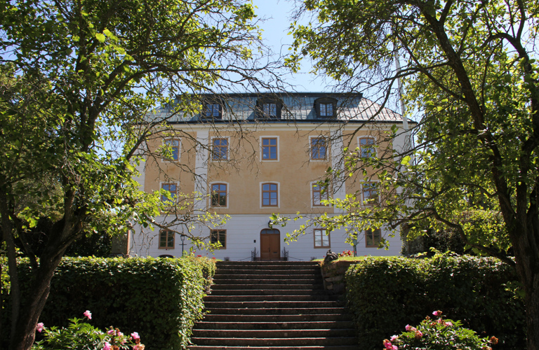 Gävle slott sett från den grönskande slottsparken. En gulputsad fyravånings-byggnad med brutet tak och tak-kupor.