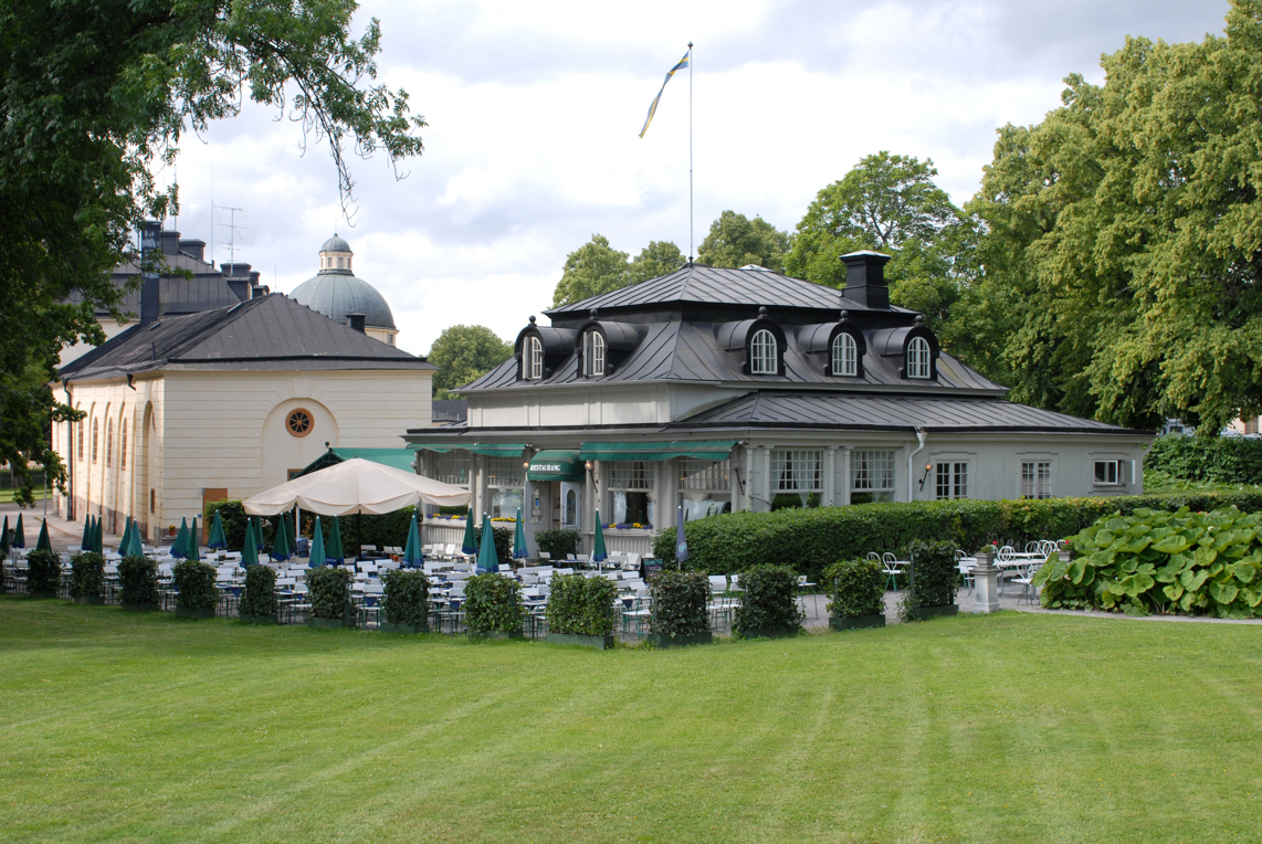 Karamellan vid Drottningholms slott. En vit-putsad byggnad med brutet tak och tak-kupor. Sommar och grönska.