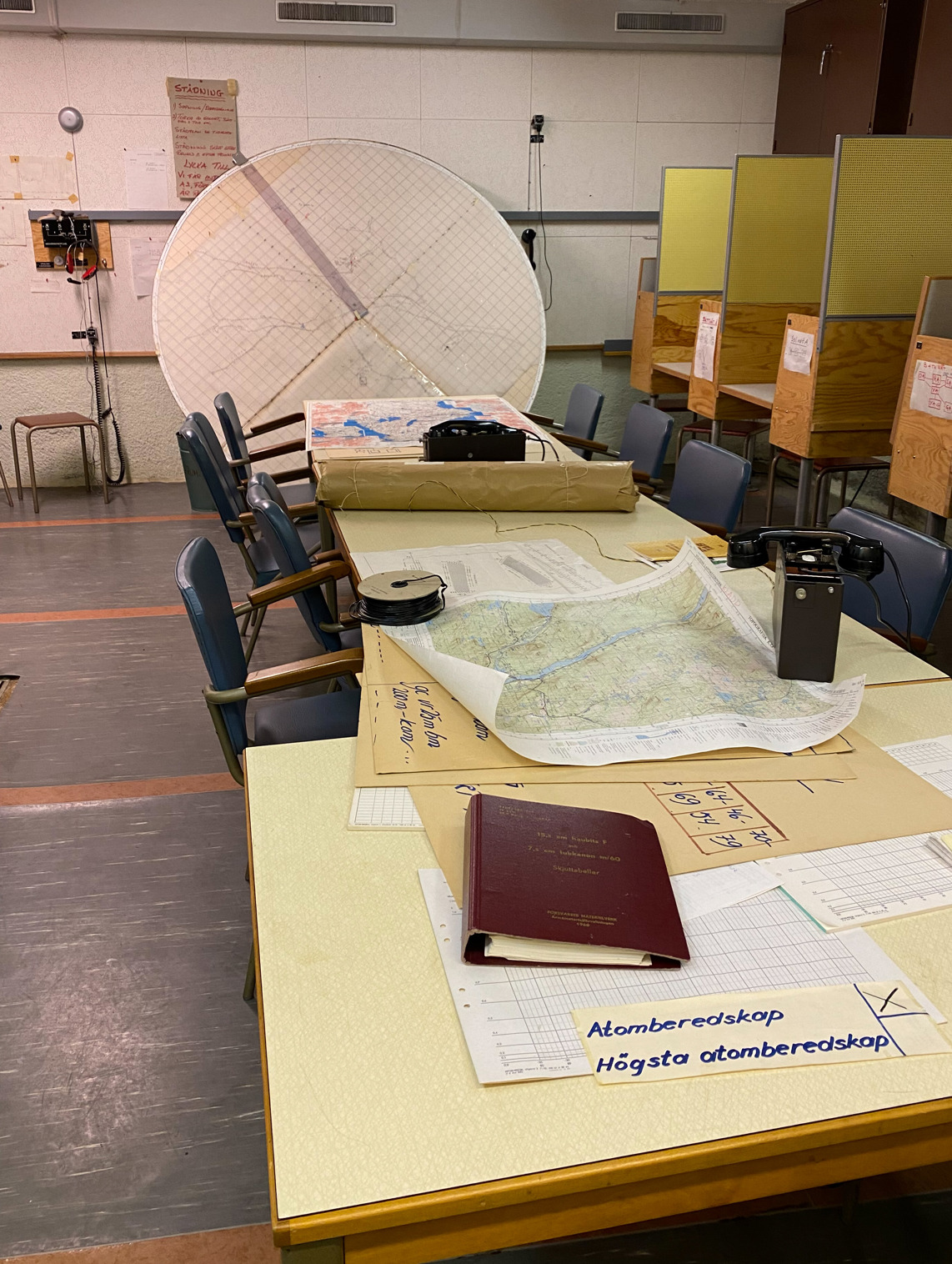 Ett rum under jord med vita väggar med djuddämpande material. Grå linoleummatta på golvet. Ett stort bord i mitten är fyllt med pappar och kartor. På en lapp står "Atomberedskap, Högsta atomberedskap". Längst bort i rummet står en stor rund karta. Till höger i bild syns en rad med bås med arbetsplatser.