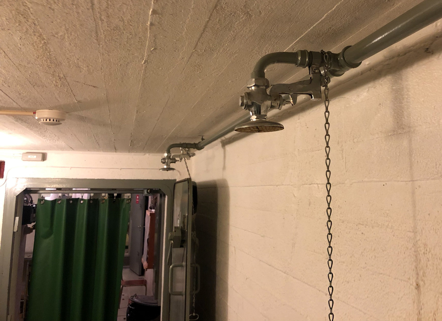 Ett genomgångrum under jord. Betongväggar målade i vitt. På bilden syns två duschmunstycken av metall fästa uppe en vattenledning vid taket och framför en dörr anas ett tjockt grönt gummidraperi.