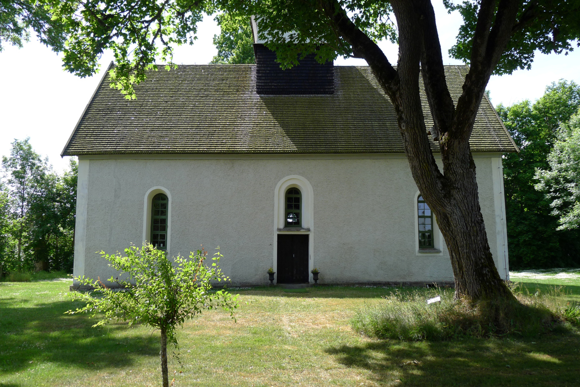 Brunneby kyrka. En liten, vitputsad medeltida kyrka med rundbågiga fönster. Sommar och grönska.