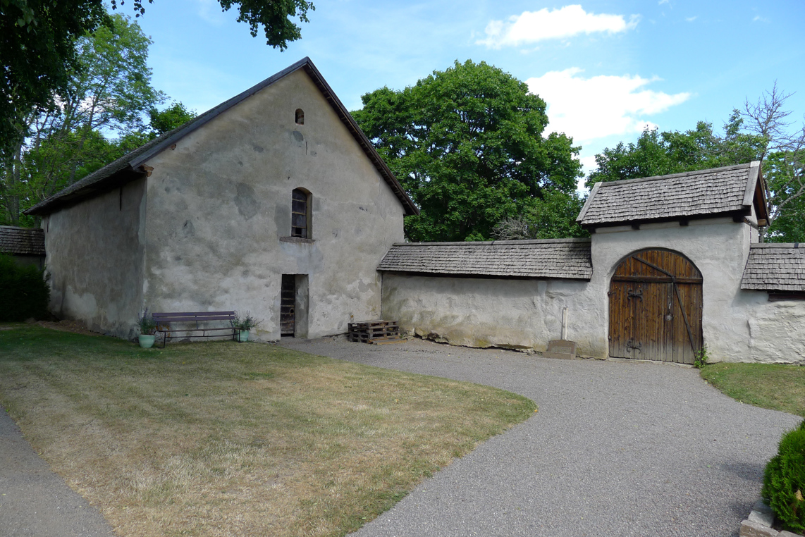Medeltida stenbod vid muren kring Risinge gamla kyrka. Muren och boden är vitputsade och har spåntak. I muren sitter en träport.